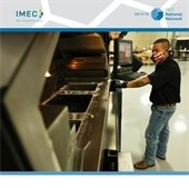 A masked manufacturing worker in a black polo and jeans works on a metal industrial sink.