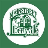 A white circle is on a dark green background. Inside the circle is a logo of a historic house and tree in between the words "Mainstreet Libertyville."