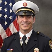 A fire chief in uniform is smiling in front of an American flag.