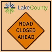 An orange diamond-shaped sign says "Road Closed Ahead" on a light orange background. A sunburst logo and "Lake County" are above in blue and green.